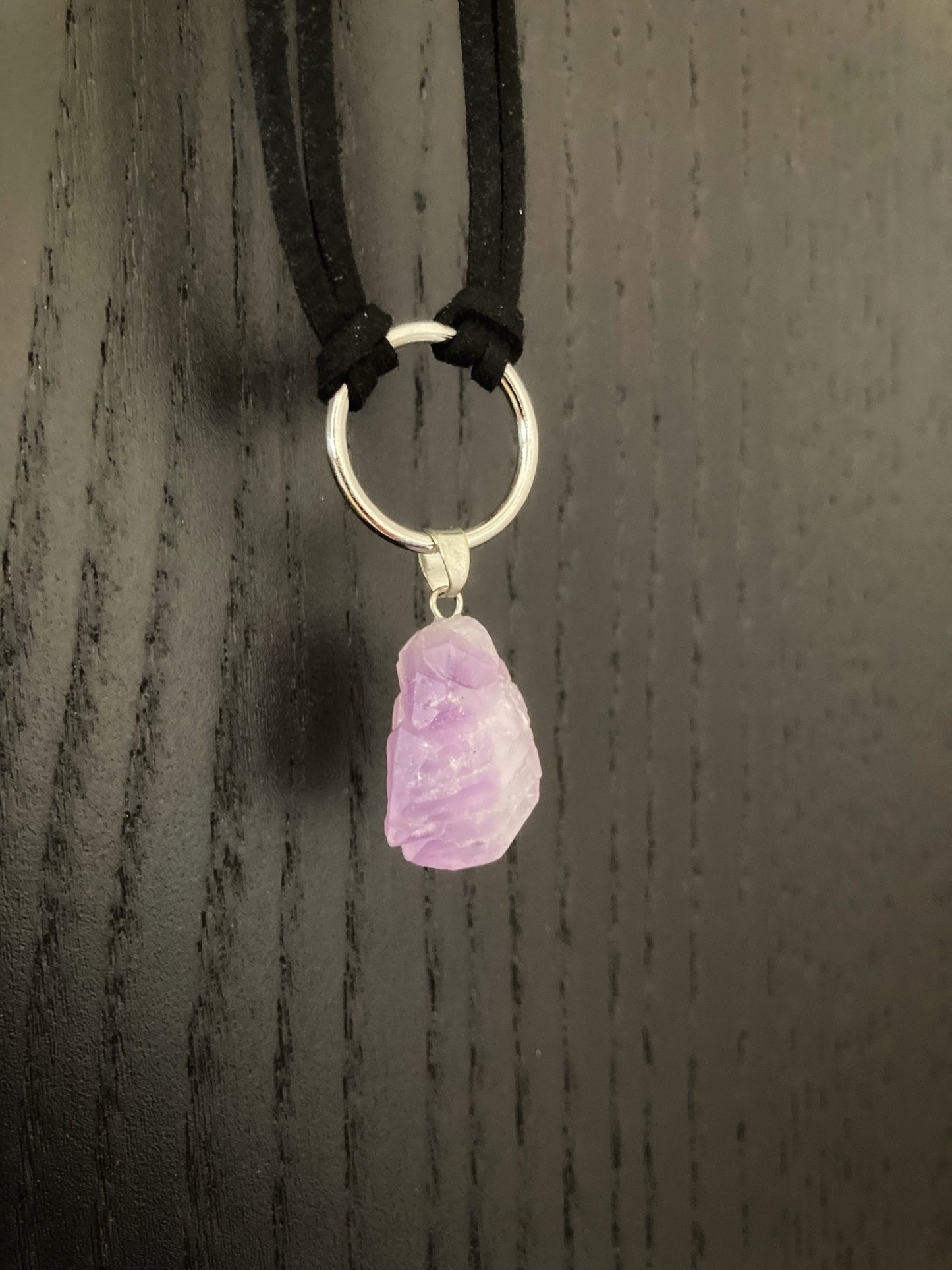 Hanging on a black background is a black cord necklace wirth a purple crystal attached to it via a metal ring., The crystal is small and rugged looking.