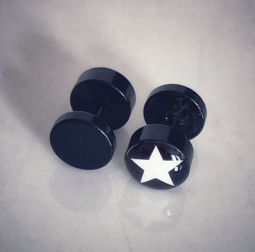 A shiny surface holds 2 black fake plug earrings. The show the opposite ends of the items with one having a white star printed on it and the other being plain.