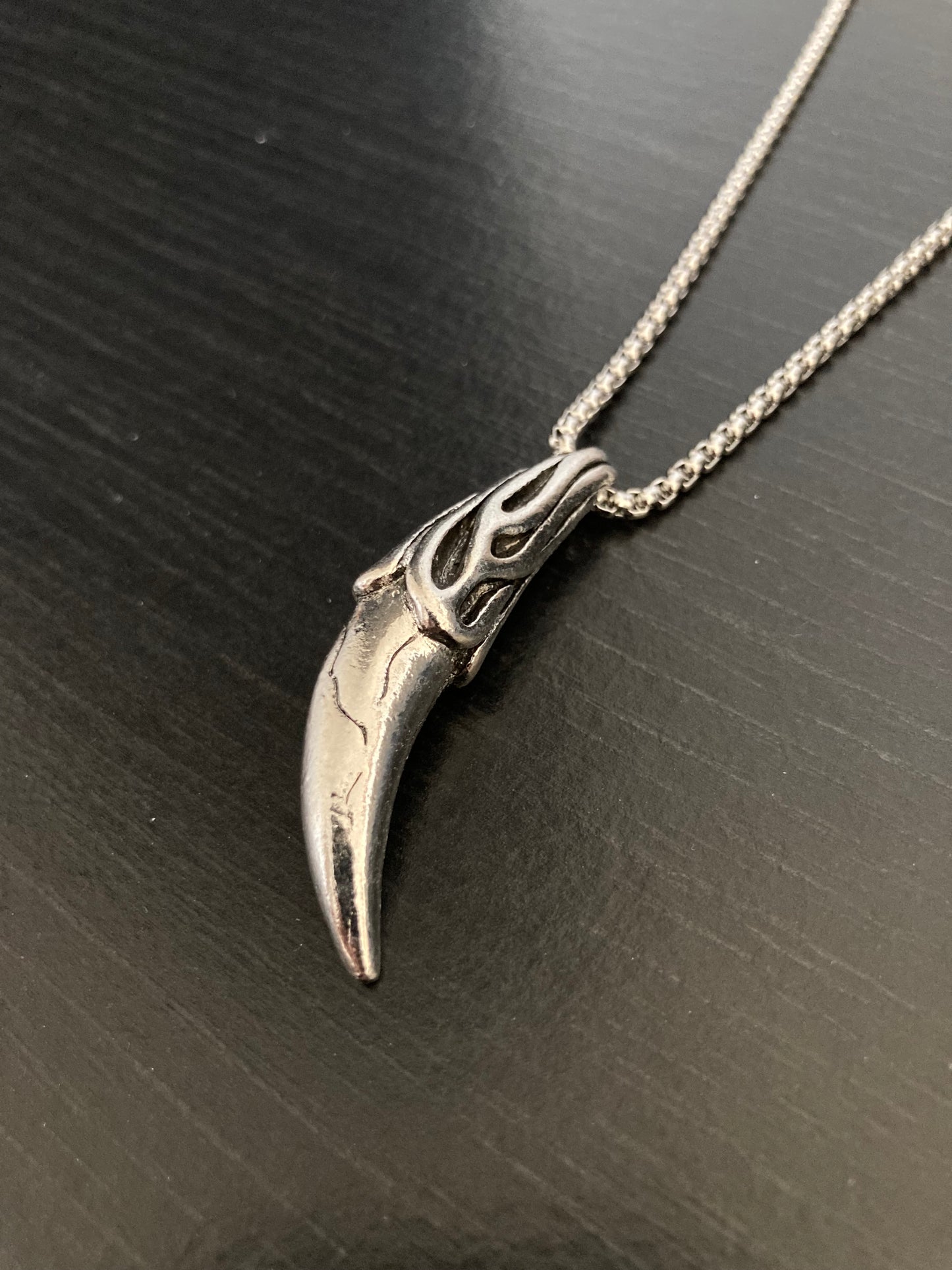 A side view of a tooth pendant lying on a grainy black background. There is a chain attached to the item which is shaped like a wolf's tooth with intricate detailing along the length of it. It is shiny to look at and solid to hold in your hand.