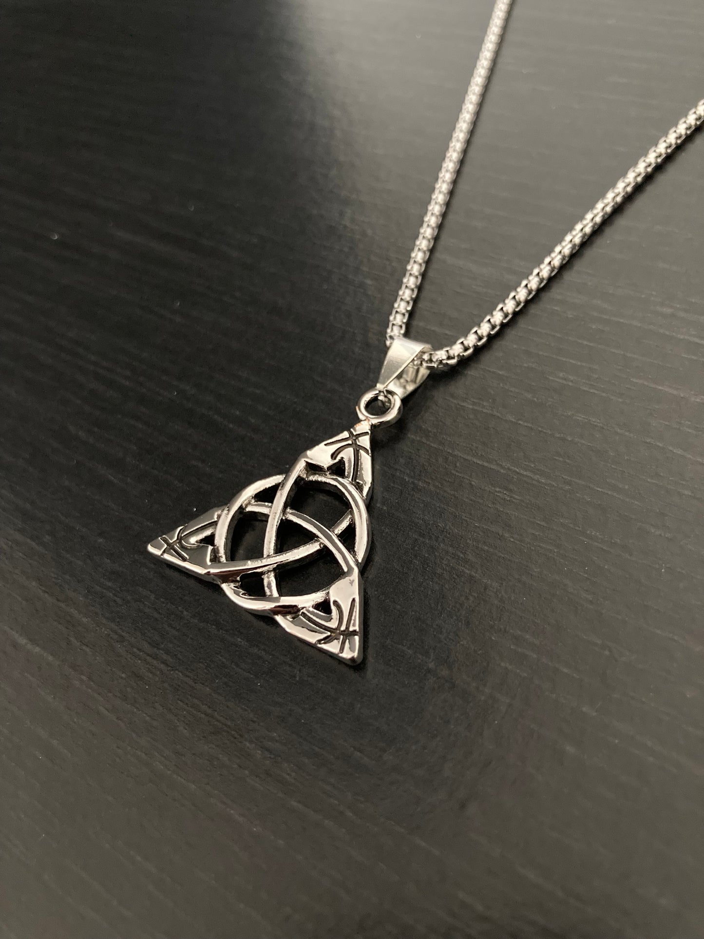 An angled view of a triangular shaped pendant attached to a chain, both silver in colour and made of stainless steel. This necklace is lightweight to hold and wear but of high quality still. There are interlocked circles in the middle of the pendant.