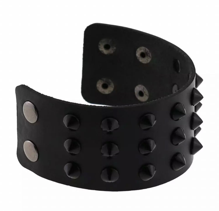 A view of a bracelet that is sitting undone on a white surface. It is black and is very wide. There are spikes attached to it all over it and there are 2 silver coloured press studs for fastening with a couple of different size options making it adjustable.