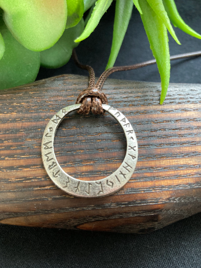 Lying on a  wooden item is a stainless steel round hollow pendant attached to a brown cord necklace. There are rune symbols engraved onto the outer egde and the whole thing feels lightweight to hold but also quality.