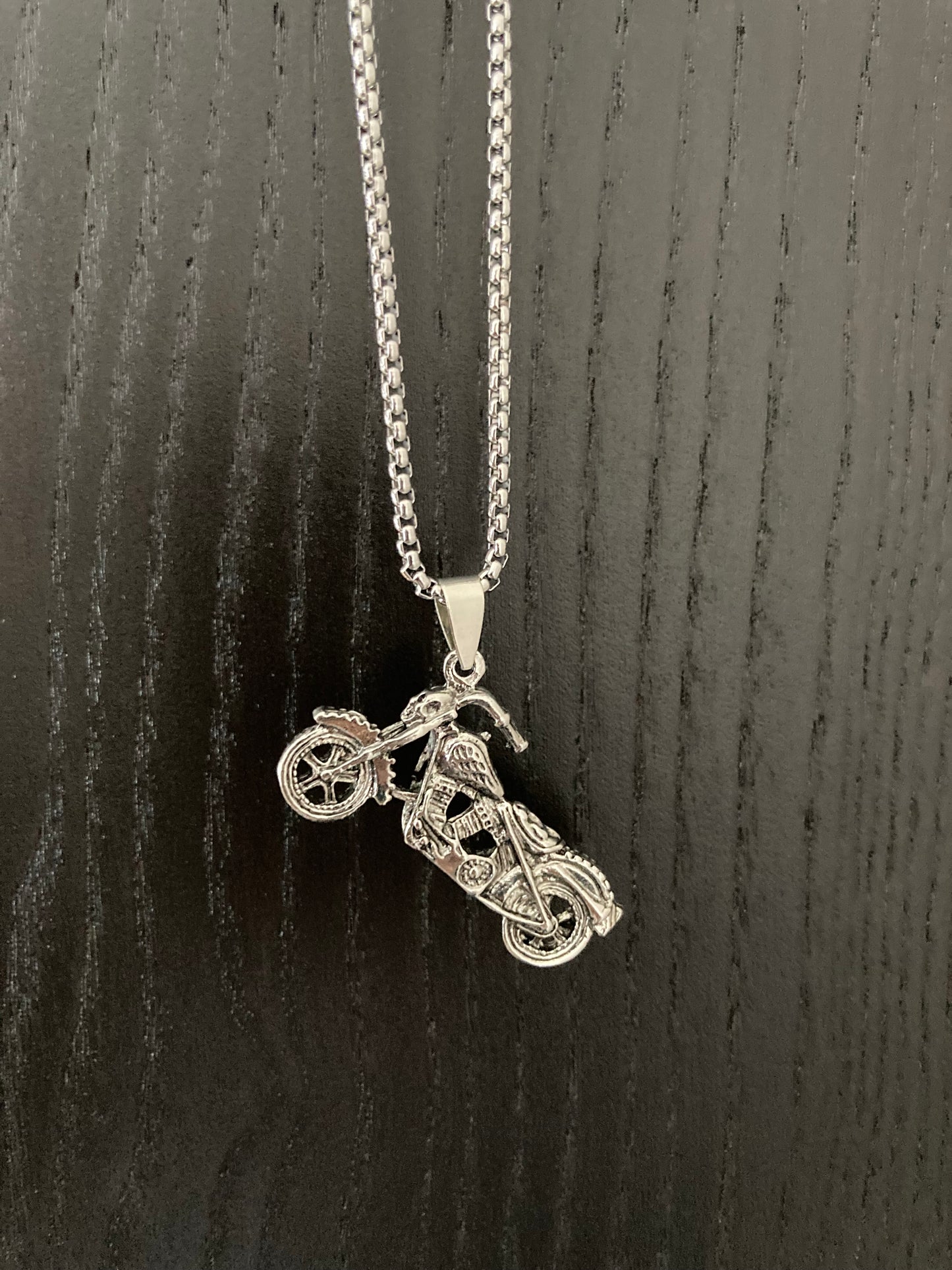 On a  grainy black surface is an angled view of a motorbike necklace. The chain looks to be quality and sturdy. The pendant itself is in the shape of a harley style bike and has real intricate markings on it.