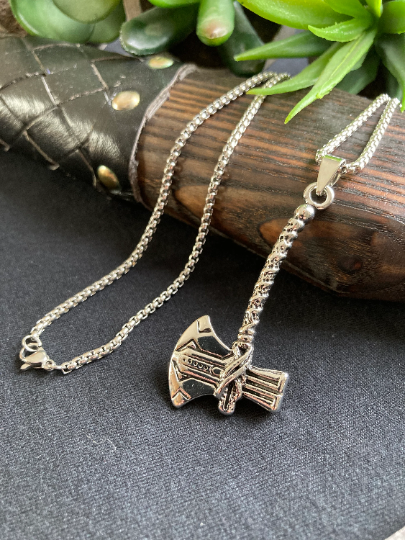 A wooden axe handle with leather on it sits on a grey surface with a stainless steel necklace draped over it. Attached to the chain is a pendant in the shape of axe with a long handle and both of these are silver in colour. 