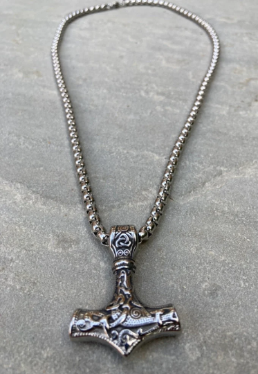 A stainless steel chain with a solid pendant in the shape of a viking hammer sits on a grey background. You can see the metal clasp and the chunkiness of the whole item. The main part of the hammer features a textured design that is stunning.