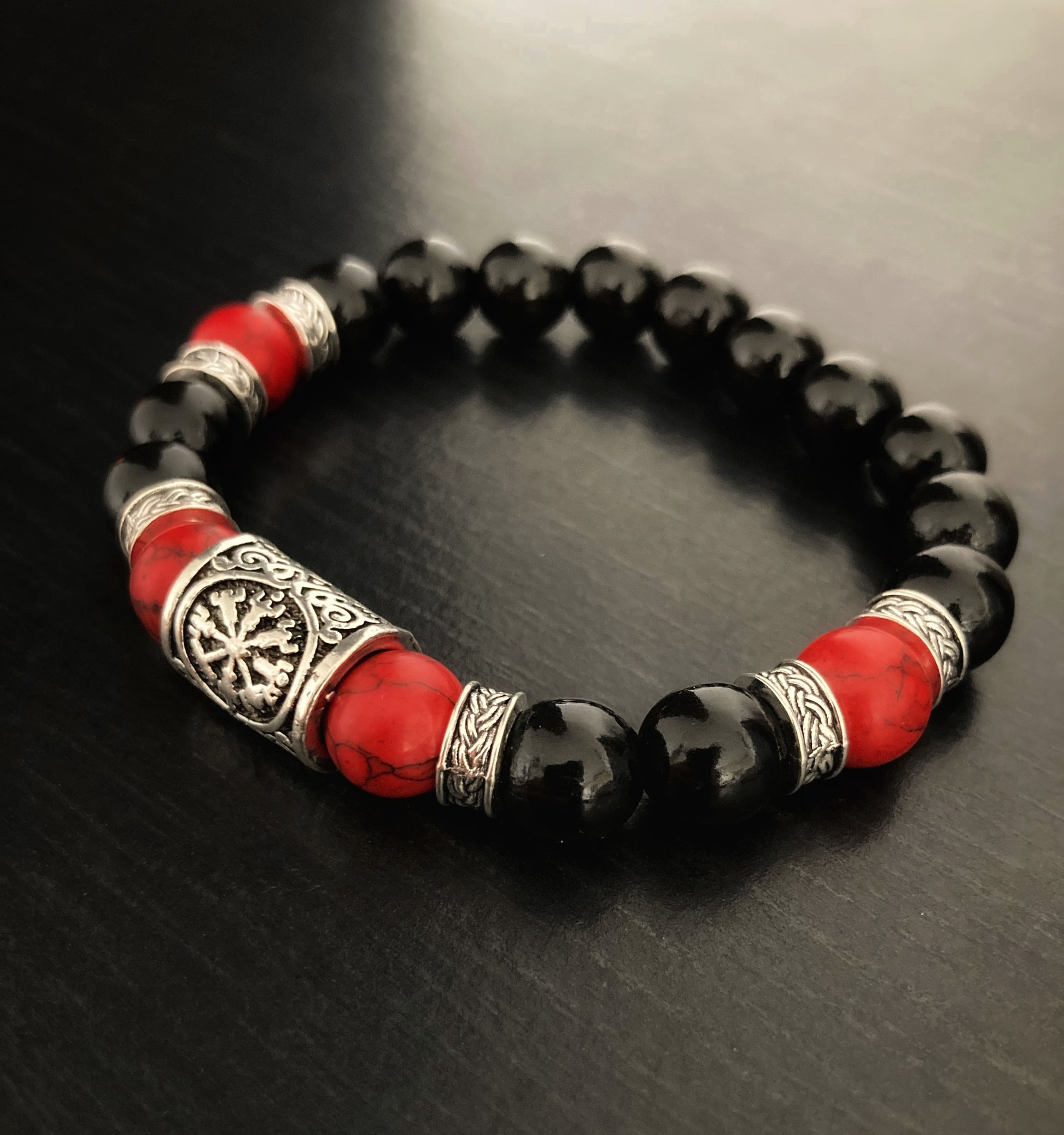 A side view of a beautiful handmade bracelet. With a silver rune bead as the main piece this is then surrounded by both black beads and red turquoise healing beads. There are also metal patterned pieces interspersed throughout and it is stunning.