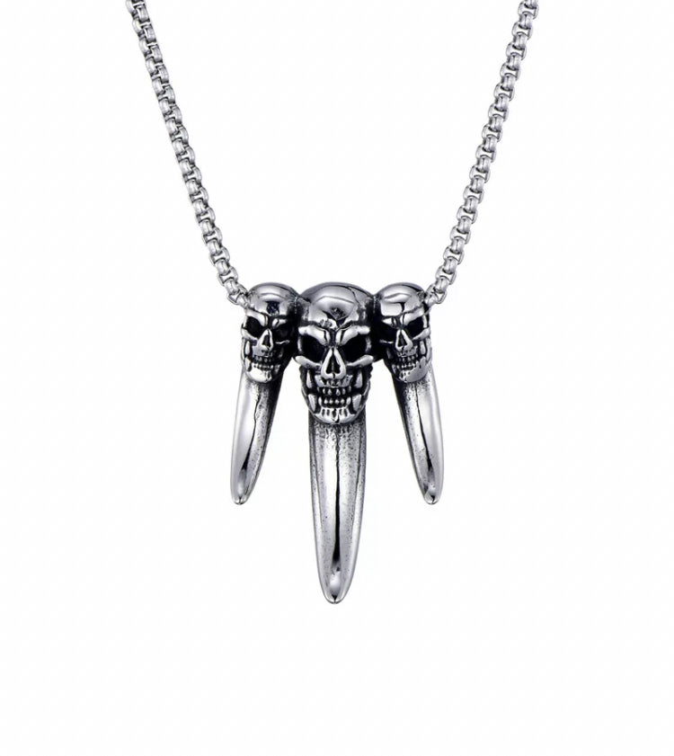 On a white background hangs a pendant which consists of three fangs next to each other all of which have skulls engraved onto the top of them. It is silver in colour and the middle fang is slightly longer than the other two which are the same length.