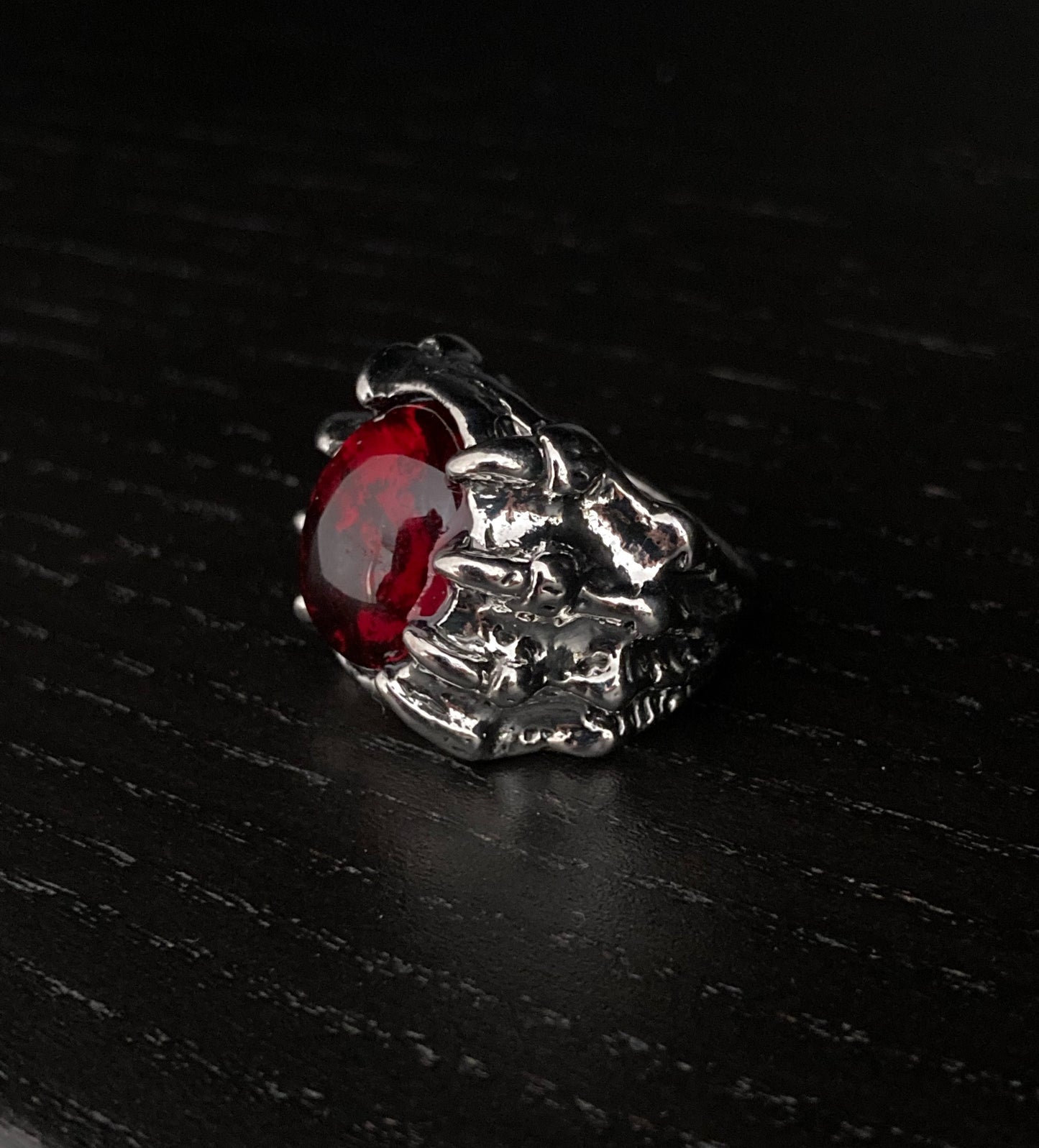 A silver coloured ring sits at an angle on a grainy black surface. It is made to look like a dragon claw that is holding onto a red stone which is made of glass. The claws have detailed engravings on them making them look realistic.