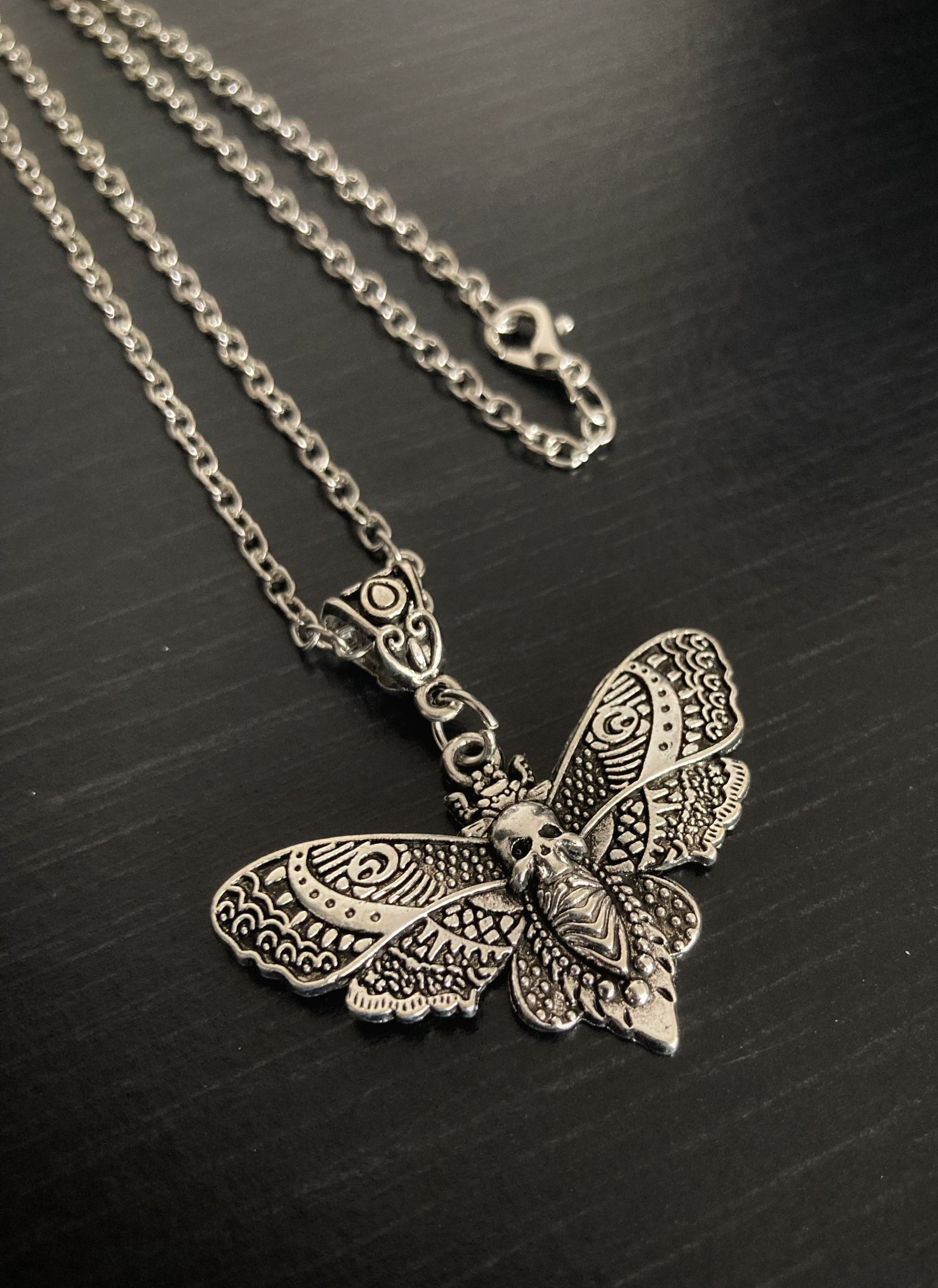 An angled view of a silver coloured chain and pendant featuring a metal clasp. The design of the pendant is in the shape of a death moth with the wings featuring a very detailed pattern. 