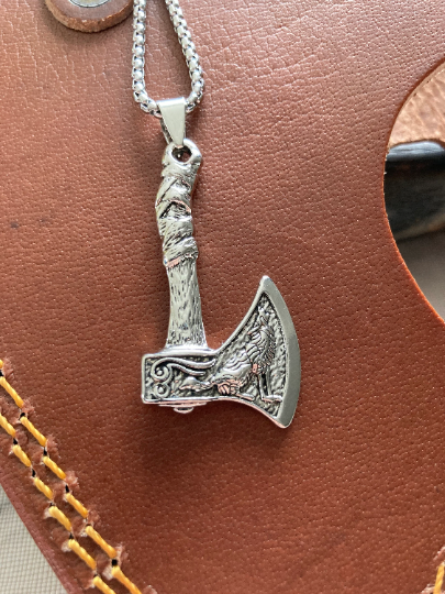 A stainless steel axe style pendant with chain sits on a brown leather item. You can see the detailing of the wolf design and feel it when you touch it. The item is shiny silver coloured to look at as is the chain. 