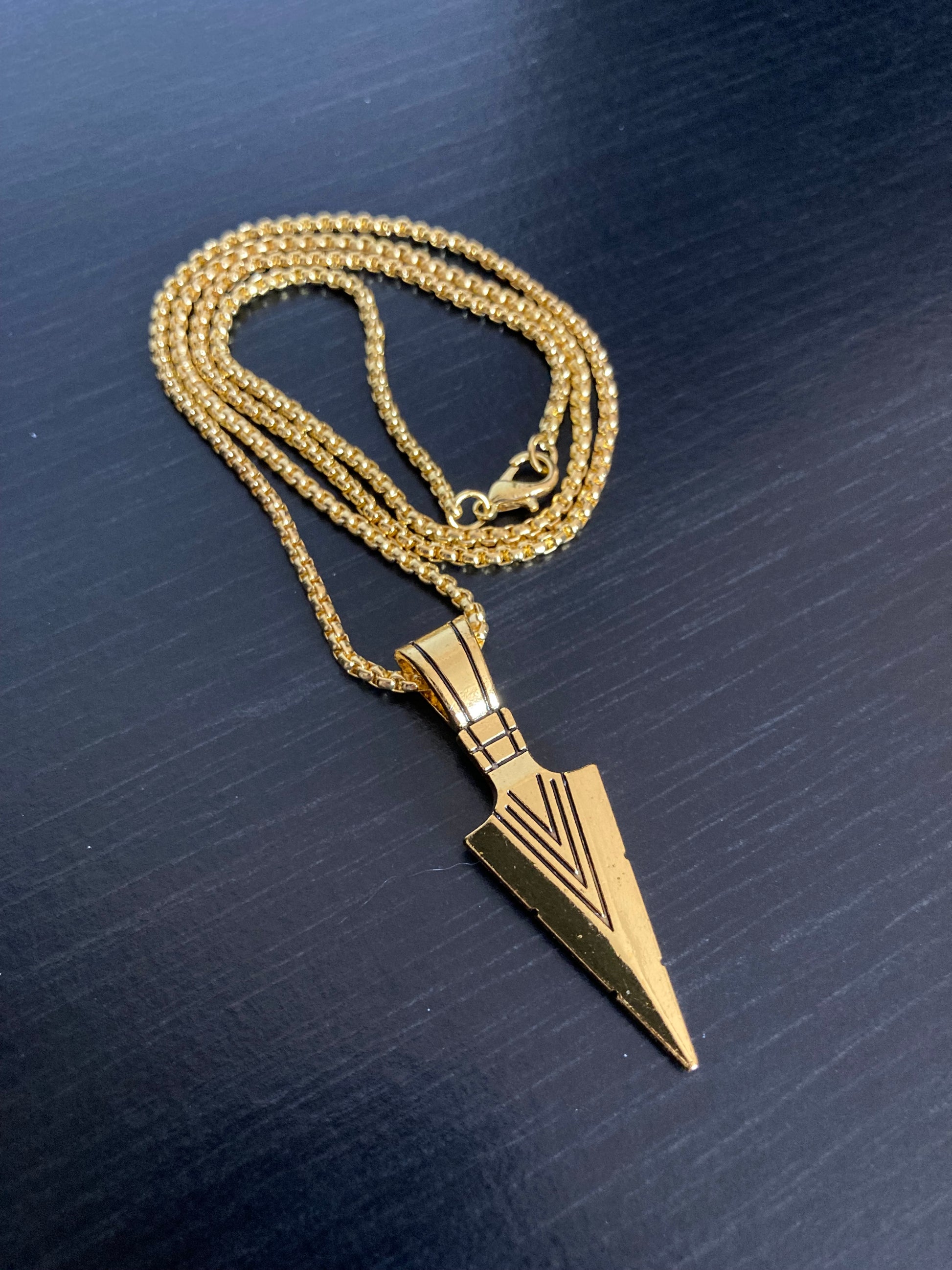 A curled up chain with a metal fastening clasp is on show here, it is attached to a arrow head pendant and both are gold in colour. The markings on the arrow stand out and look stylish as well as cool.