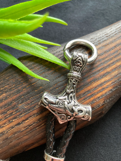 A close up of the hammer part of a bracelet is shown to be sitting on a wooden surface with a green plant in the background. This is made of stainless steel and is beautiful in its patterned detailing and silver in colour.