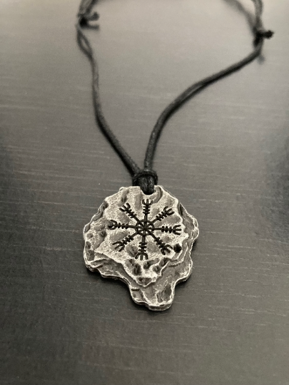 A close up of a helm of awe inscribed rock pendant on a grey textured background. This item is shiny to look at and is very solid and weighty. Looking like an actual rock this isnt defined in shape and is rugged looking and silver in colour. 