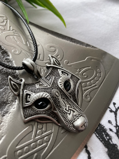 A close up view of the wolf's head pendant that is featured here lying on an axe blade at an angle. The stunning detail including the all the engravings on the face are on display and can also be felt when touching the item. 