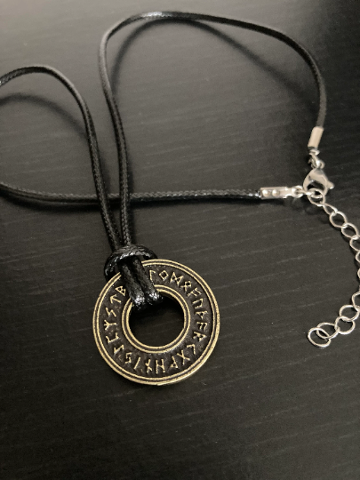 A black cord type necklace lies on a black surface. It has a metal clasp for fastening and tied to it is a disc shape with rune symbols engraved in it.