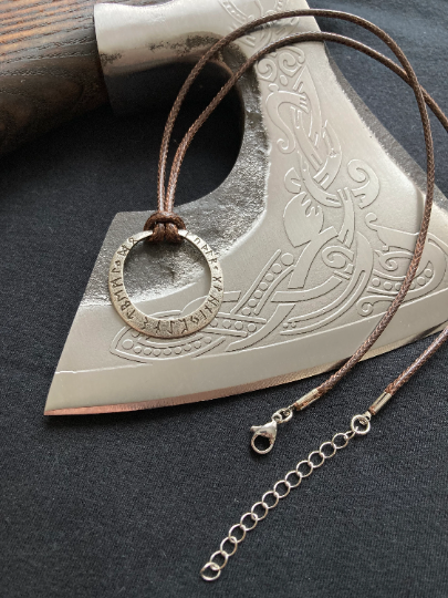 A view from above of an axe blade with a brown corded necklace draped over it. Attached to the cord is a silver coloured rune inscribed hollow circular pendant which is lightweight stainless steel. The solid metal clasp for fastening can also be seen