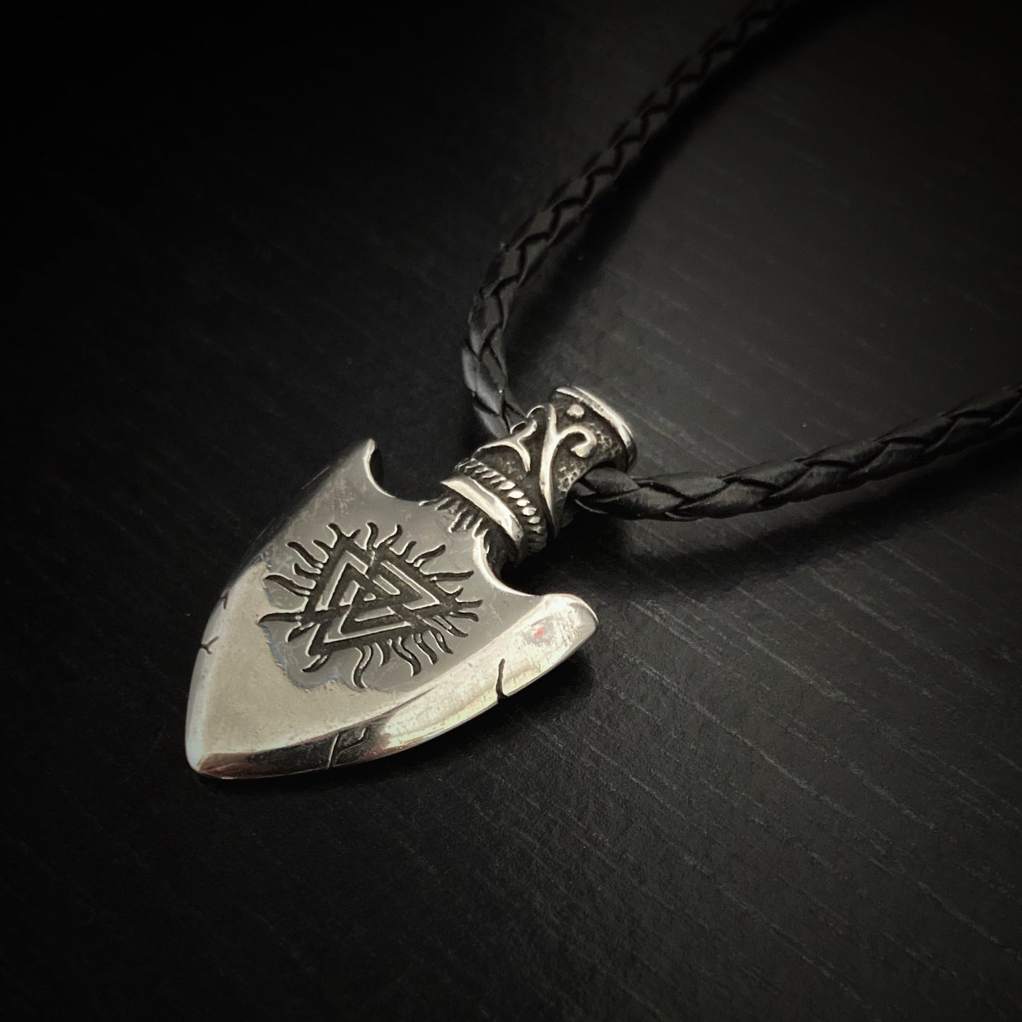 An angled view of a valknut design etched onto a silver coloured solid looking arrow head pendant. With small crack looking designs marked on it and lines coming away from the main design making it look like the edges of a sun.