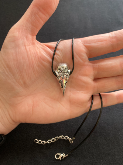 Sitting in the palm of a hand with the black cord necklace dangling beneath is a raven bird skull viking pendant. Made of stainless steel and featuring a design of the helm of awe carved onto it the pendant is lightweight but good quality.