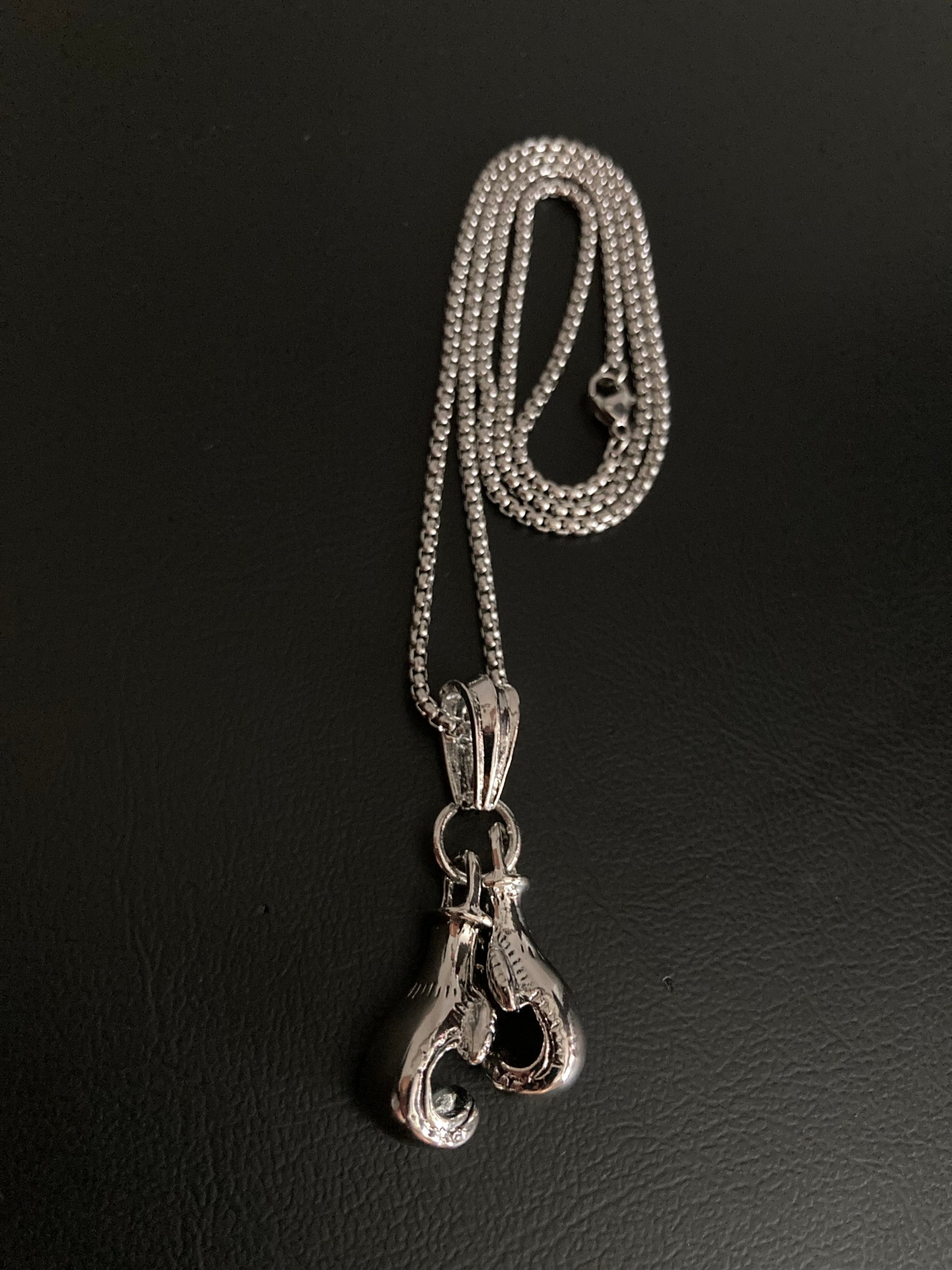 A curled up sturdy looking chain with a metal clasp sits upon a black surface. Attached to this via a ring is a pair of boxing gloves which are solid and smooth. The whole thing is silver coloured and made of stainless steel.