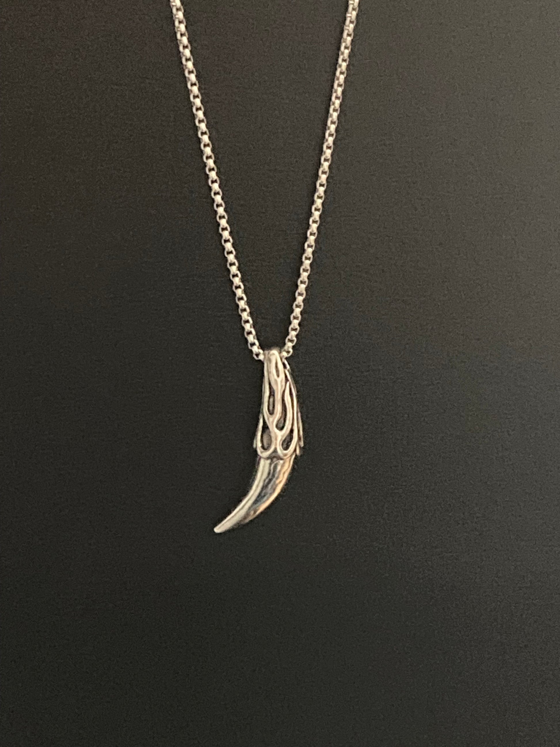 A silver coloured pendant hangs on a plain black background with a necklace chain attached to it. The item is in the shape of an animals tooth such as a wolf and is made of stainless steel. The quality of this is evident when you touch and hold it.