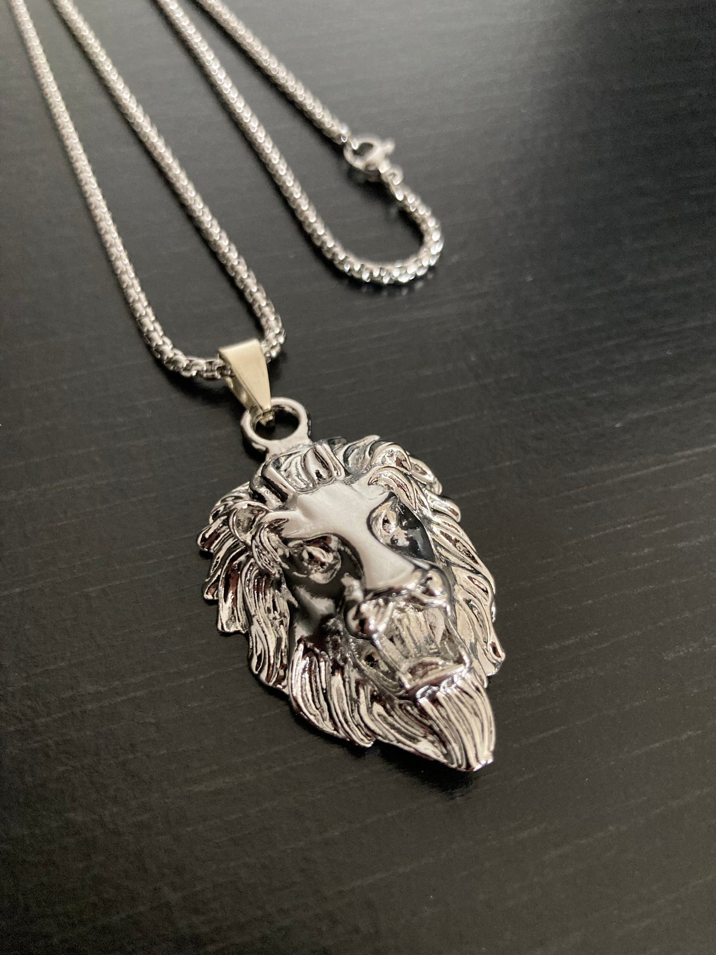 A view from above of a lion head pendant made of stainless steel. You can see the chain attached it with its metal clasp for fastening. Both are silver in colour and the lion itself has detailed features.