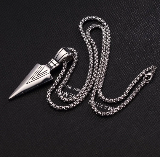 A curled up chain sits on a black background with the metal clasp in view. Attached to it is a silver coloured arrow head which is beautifully detailed and smooth to the touch. 