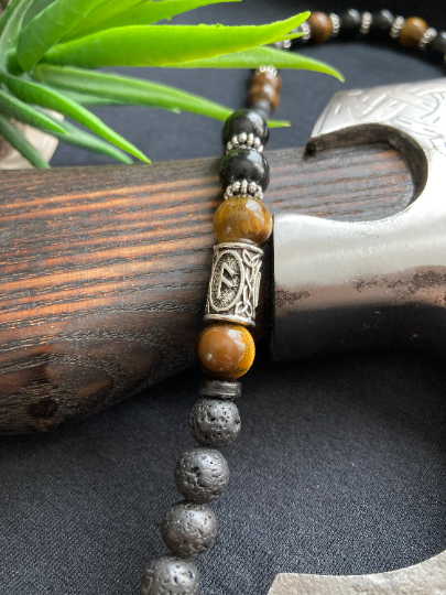 A small section of a necklace is on show and you can see textured black lava beads as well as shiny tiger eyes ones as well. There is also a silver colour metal piece with a runic symbol etched onto it.