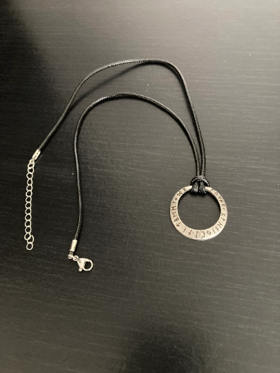 A view from above of a circular pendant upon a black background. The item is layed out so you can see the length of the black corded necklace with metal clasp wich is tied to the pendant. 