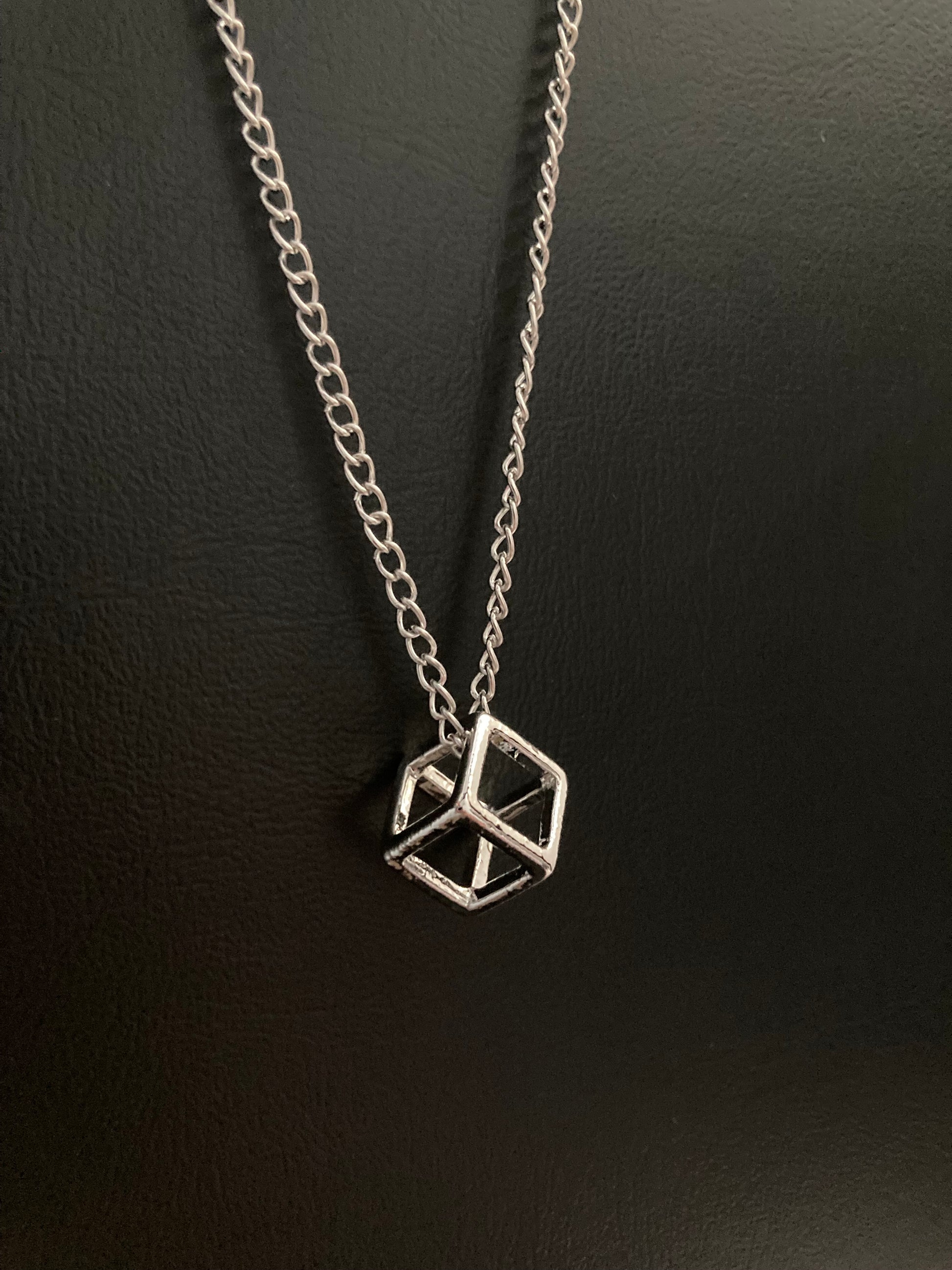 A shiny silver coloured pendant in the shape of a three dimensional cube is leaning on a black background. The hollow cube is attached to a chain and both are stainless steel.