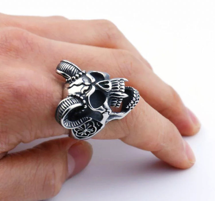 A close up of a hand with a skull ring on one finger. The item is a horned vampire with fangs and intricate markings on its face. It looks chunky and solid and is a silver colour.  