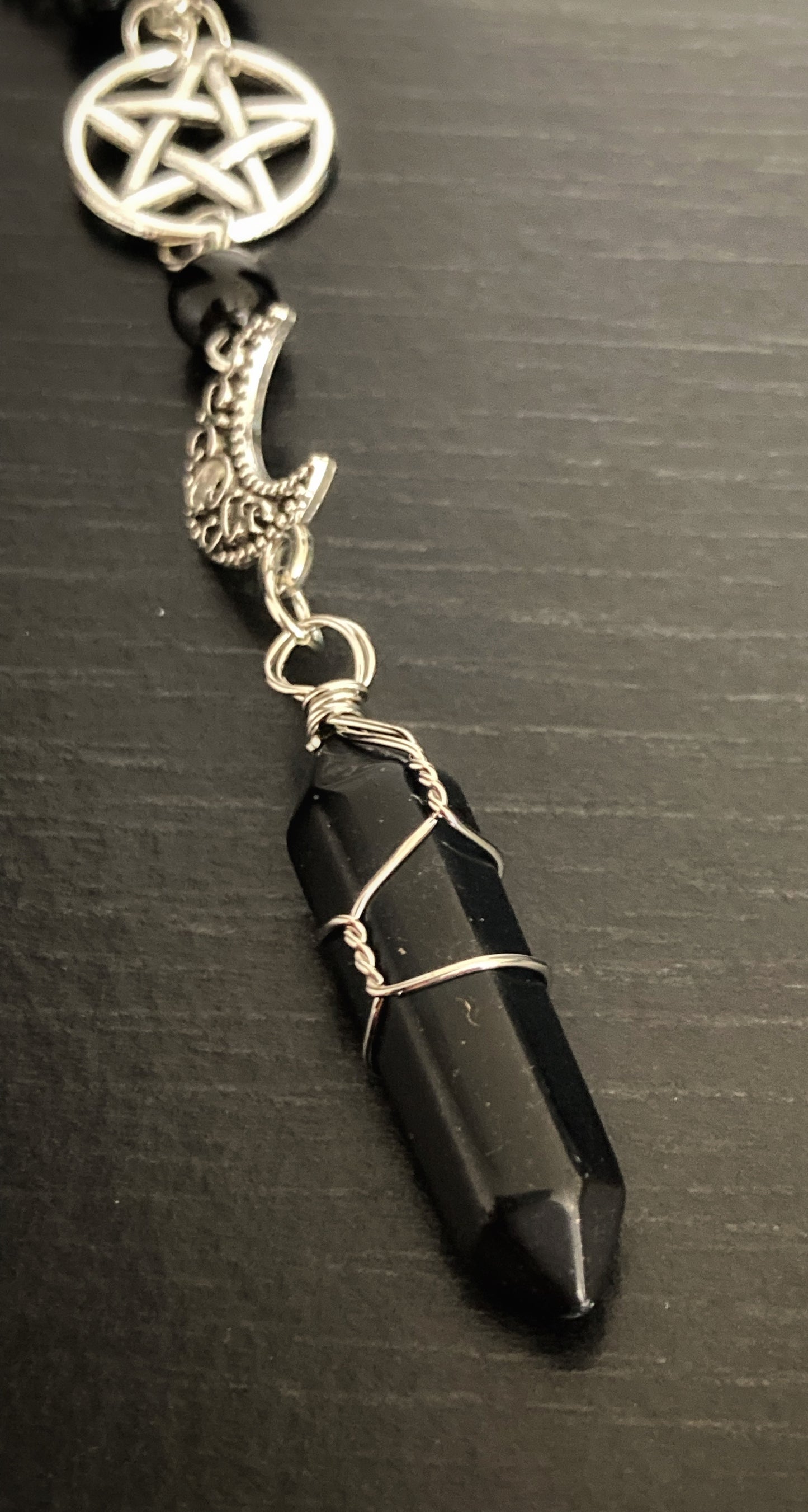 A close up view of a black onyx stone that is shiny in appearance and is held in a wire which is attached to a silver coloured crescent moon. Above that you can see a pentagram within a circle also silver in colour.