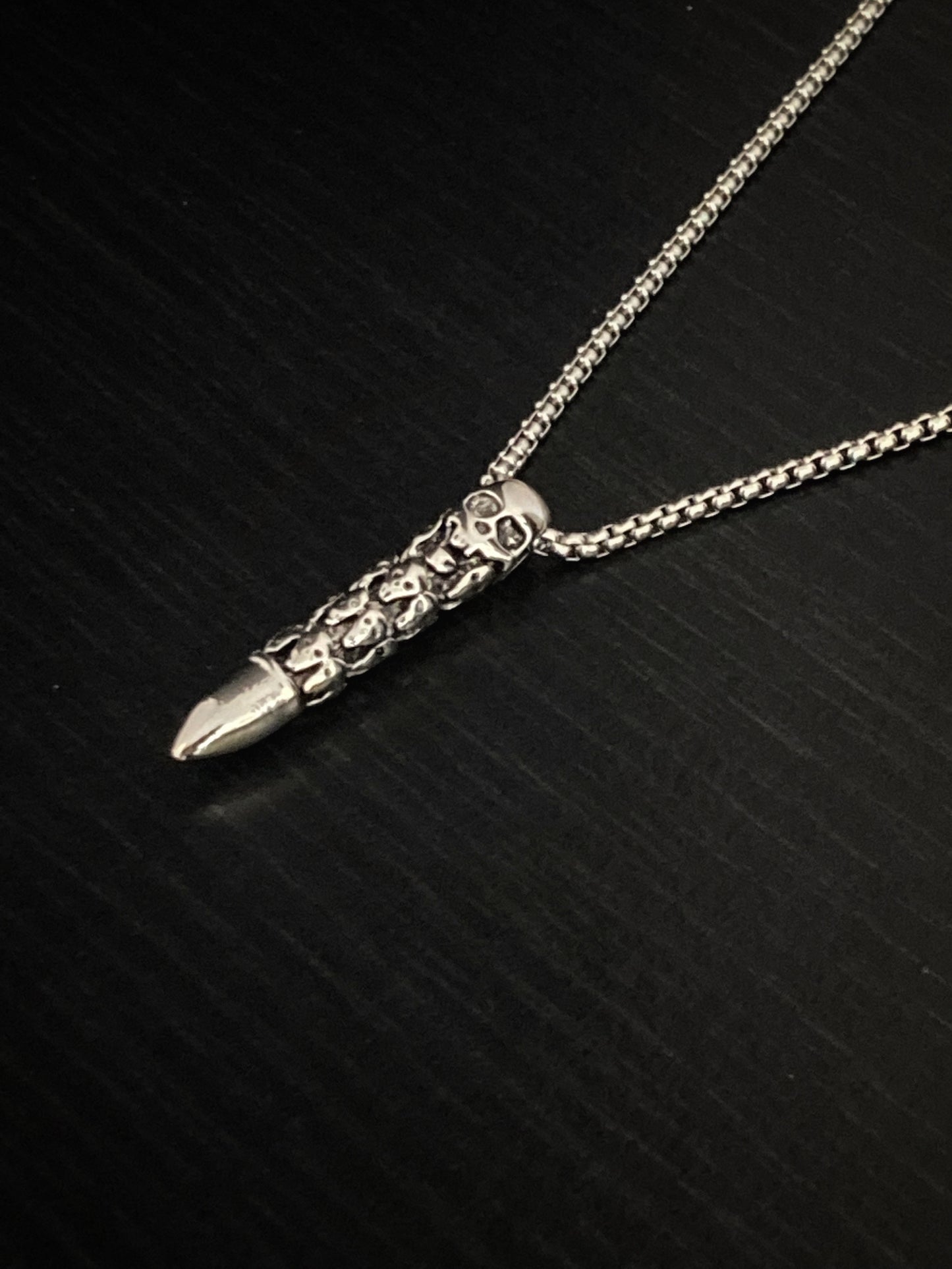 A side view of a skull carved bullet pendant on a chained necklace both of which are silver in colour and made of stainless steel. The skull heads are intricately engraved onto the main body with the tip being left plain.