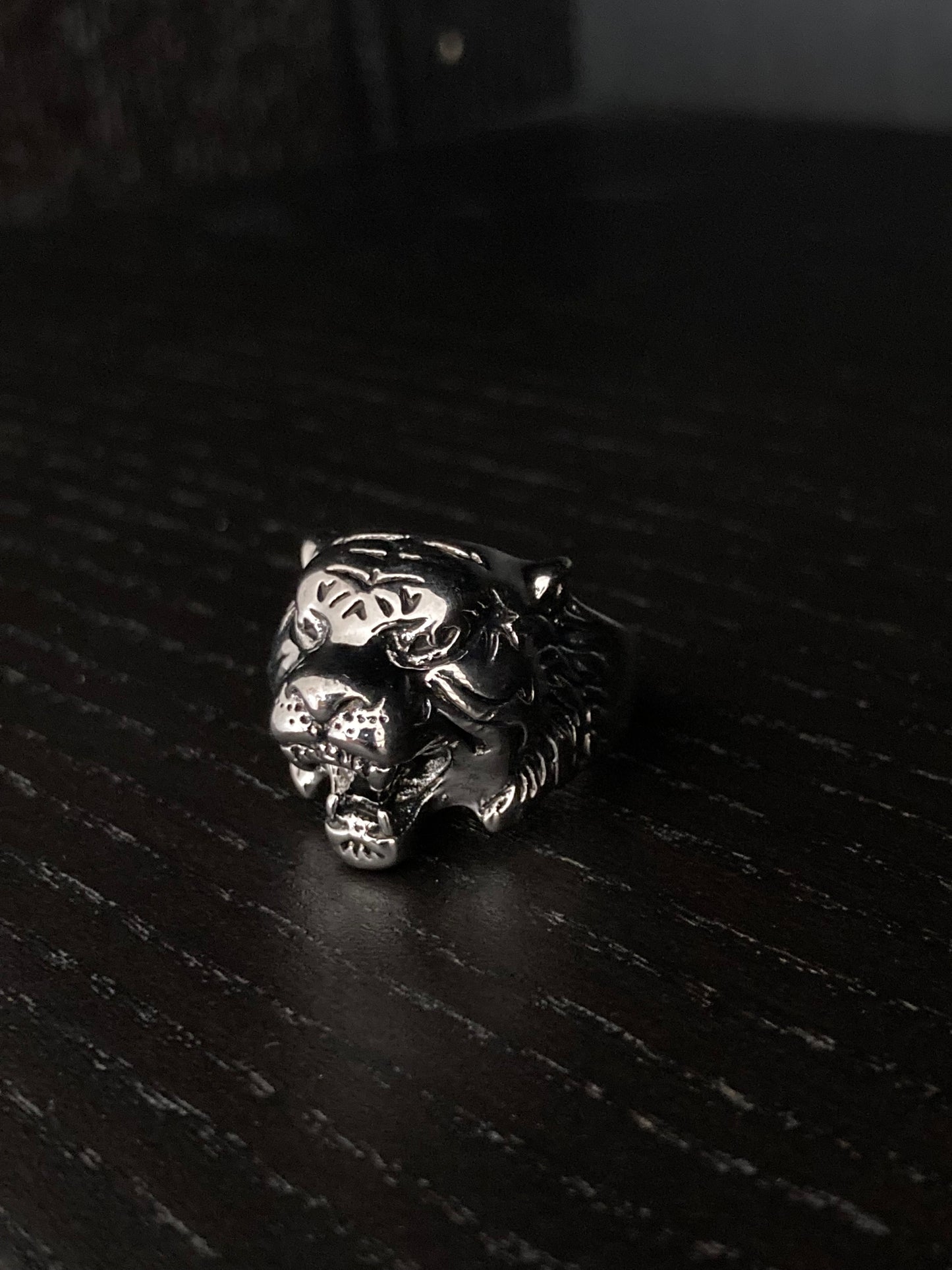 An angled view of a tiger head ring that sits upon a black grainy surface. It is ferocious looking with its mouth gaping open and the whole item is solid and weighty and shiny silver coloured in appearance. 