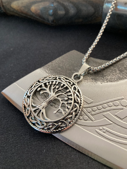 Sitting upon a viking axe blade is a silver coloured chain featuring a circular pendant that is one side in design and features the tree of life Yggdrasil. Shiny in appearance and silver in colour the tree is a cut out design.