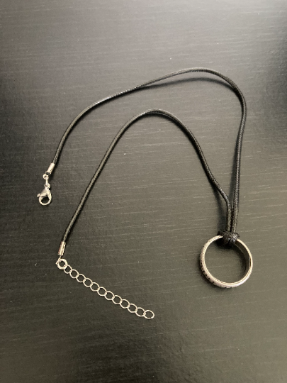 On a black background sits a ring necklace which is silver in colour with a black cord for going around the neck. You can see the metal clasp which fastens it and the length of the cord. This item is lightweight to hold but you can feel the quality.