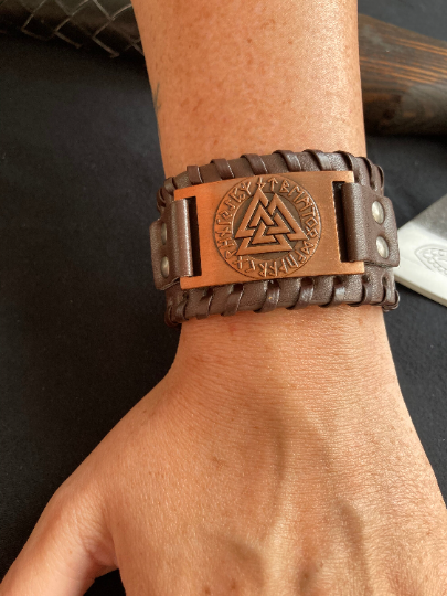 A persons wrist is showing as wearing a brown wide leather bracelet which is sitting comfortably by the wrist bone. There is a bronze metal rectangle with a design of the valknut with runes on it. The leather itself is textured on the outside.