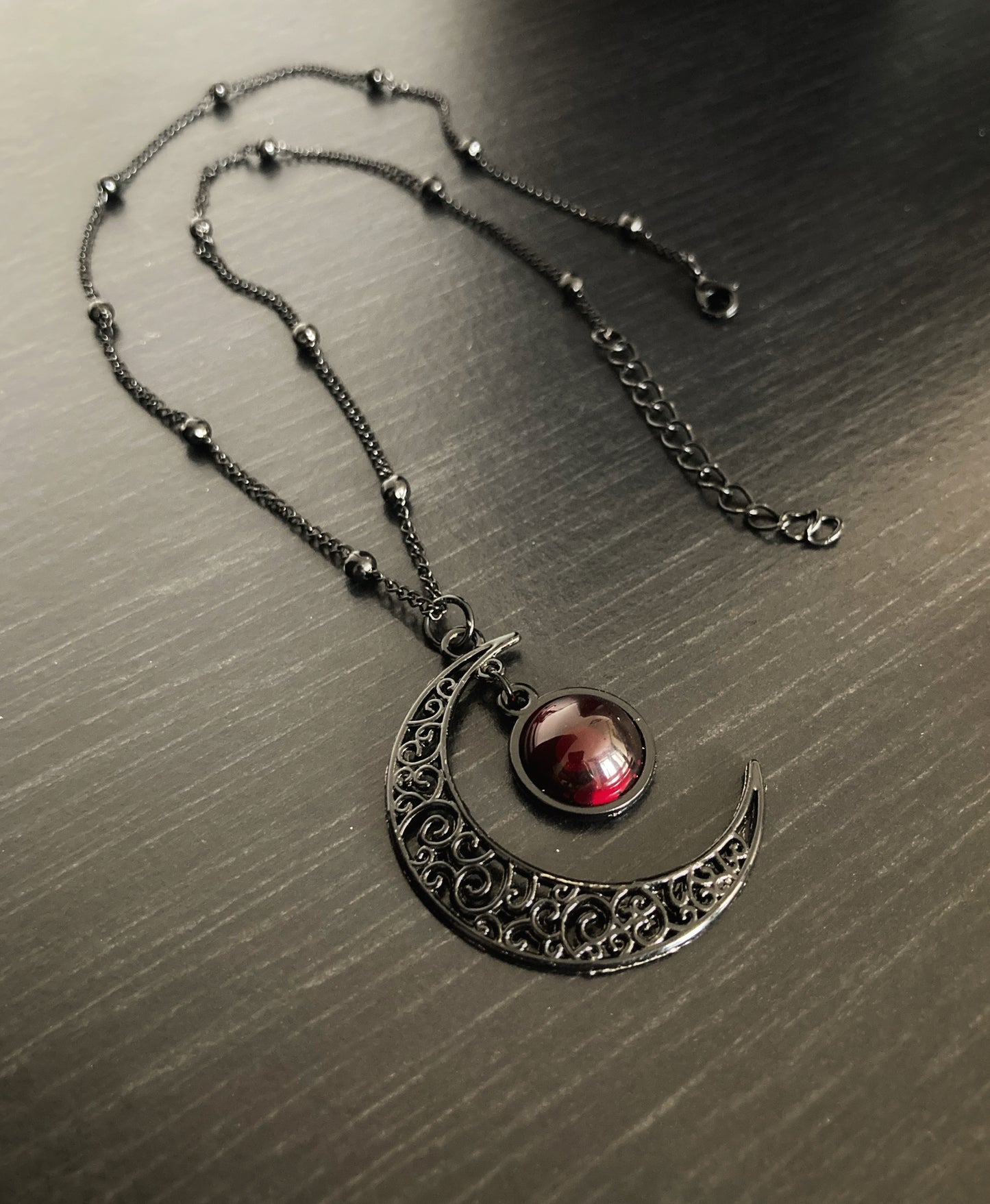 A full view of a black necklace with crescent moon pendant. You can see the metal clasp that fastens the item and periodically there are small ball shapes all along the chain.