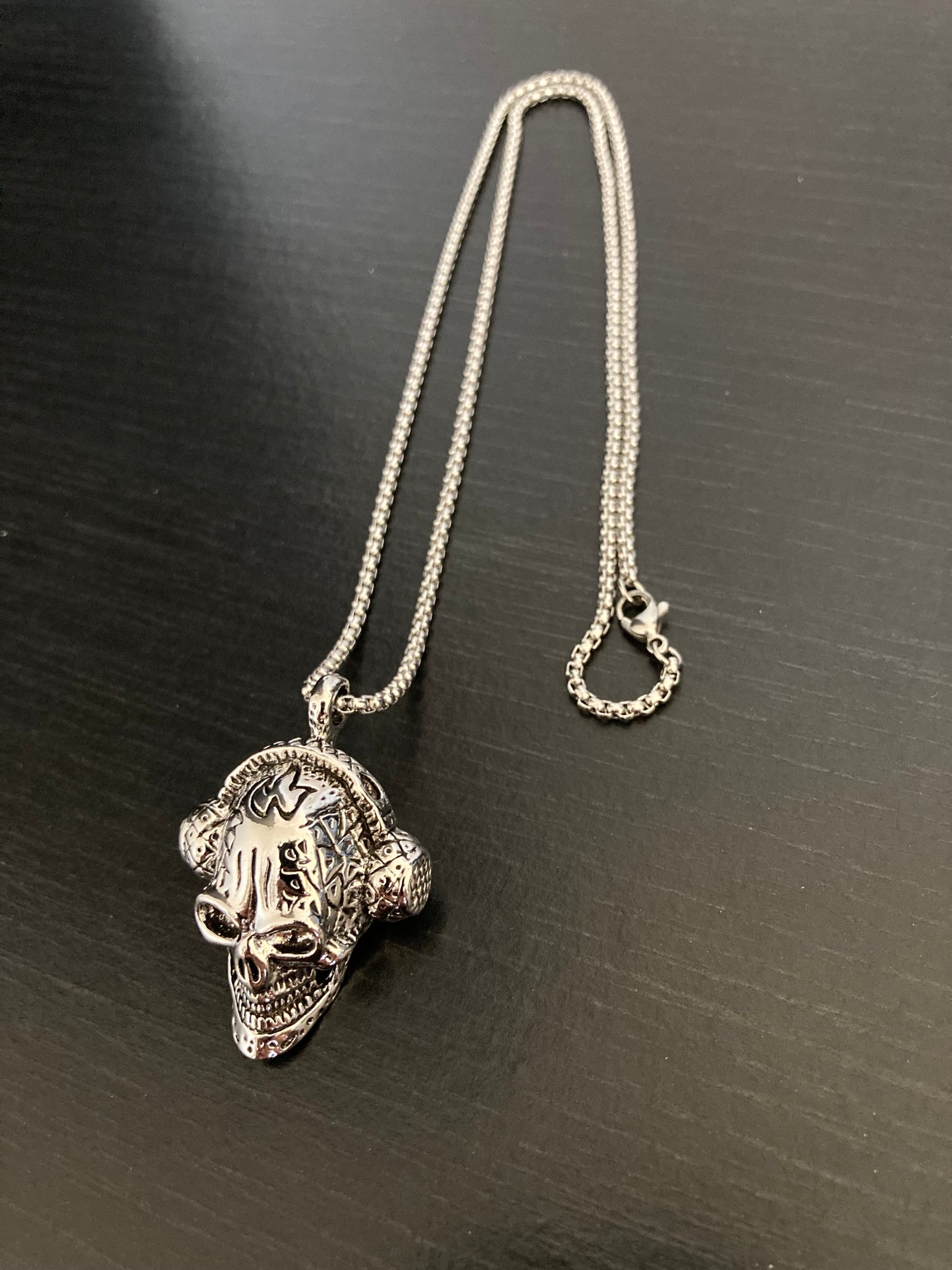 On a black background is a a view of a skull pendant attached to a study looking chain with metal clasp for fastening. Both are stainless steel and a silver colour with the skull being quite chunky in design. 