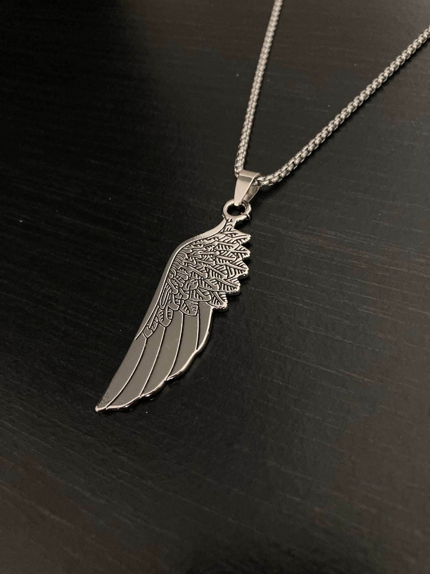 A view of a stainless steel pendant in the shape of a wing. You can see the engraved markings on the item and if you touch it you can feel the detailing. The whole necklace is light in weight with the chain feeling strong.