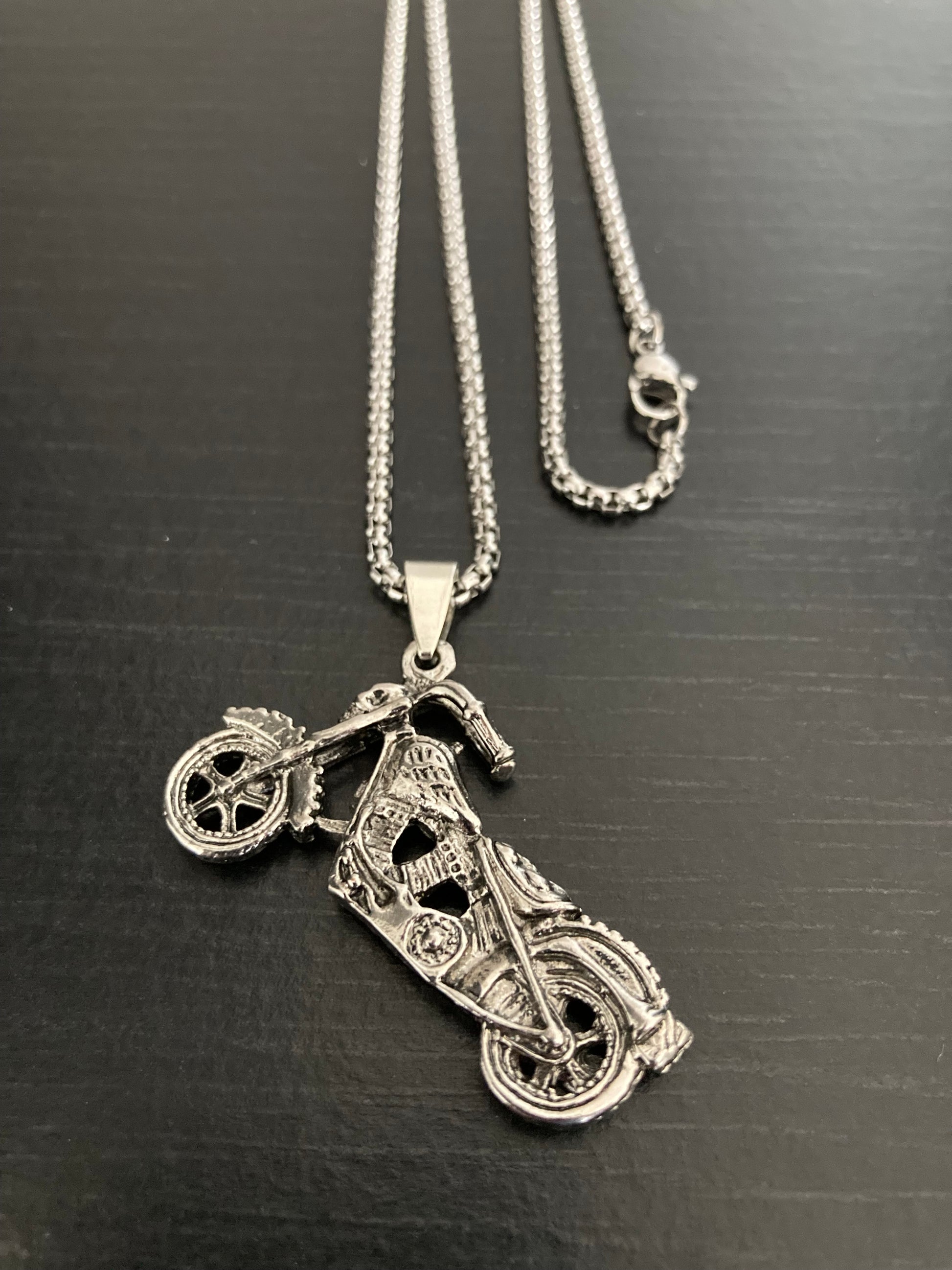 On a black surface is a motorbike necklace. The chain is showing as well as the sturdy looking clasp for fastening. The pendant itself is in the shape of a harley style bike and has real intricate markings on it.