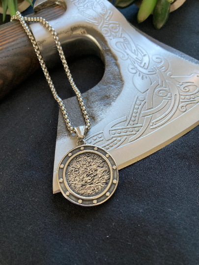 Pendant is lying on the edge of a patterned axe bladed with the chain laying on the wooden handle. The reverse of the compass is shown which is silver in colour with a swirling pattern in the middle and silver raised dots around the outside. 