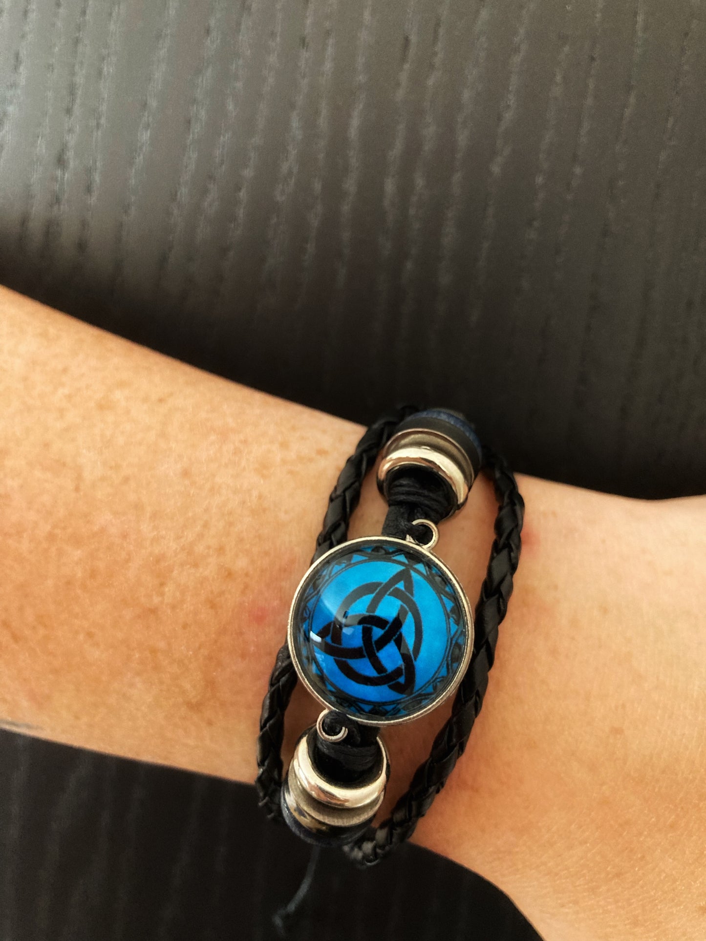 A birds eye view of a leather bracelet. You can see in detail the stunning black interlocking triangular deisgn that sits on a blue background and is covered by a clear glass lookng cover. There are beads that come off of that either side onto a solid black band. Either side of this band are two woven leather bands. 