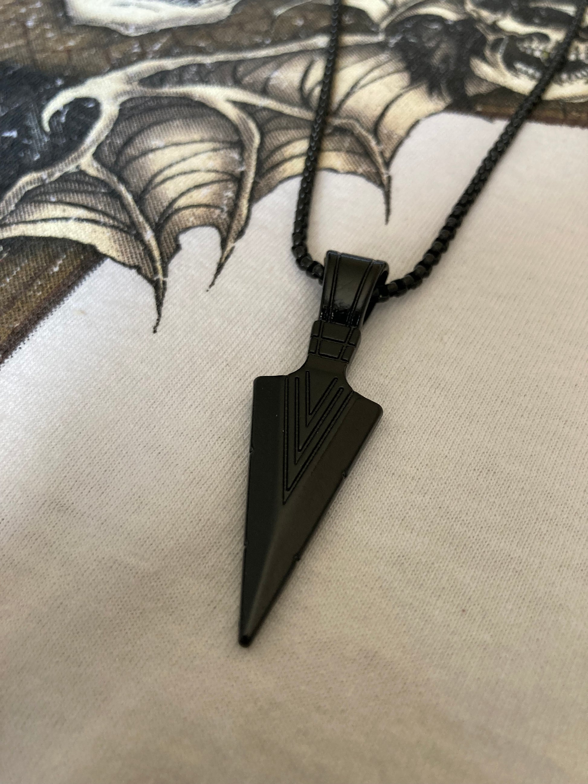 An angled view of a black chain which holds a black arrow head pendant which is both lightweight but quality. There are v shaped patterned lines on the arrow itself which ar also black.