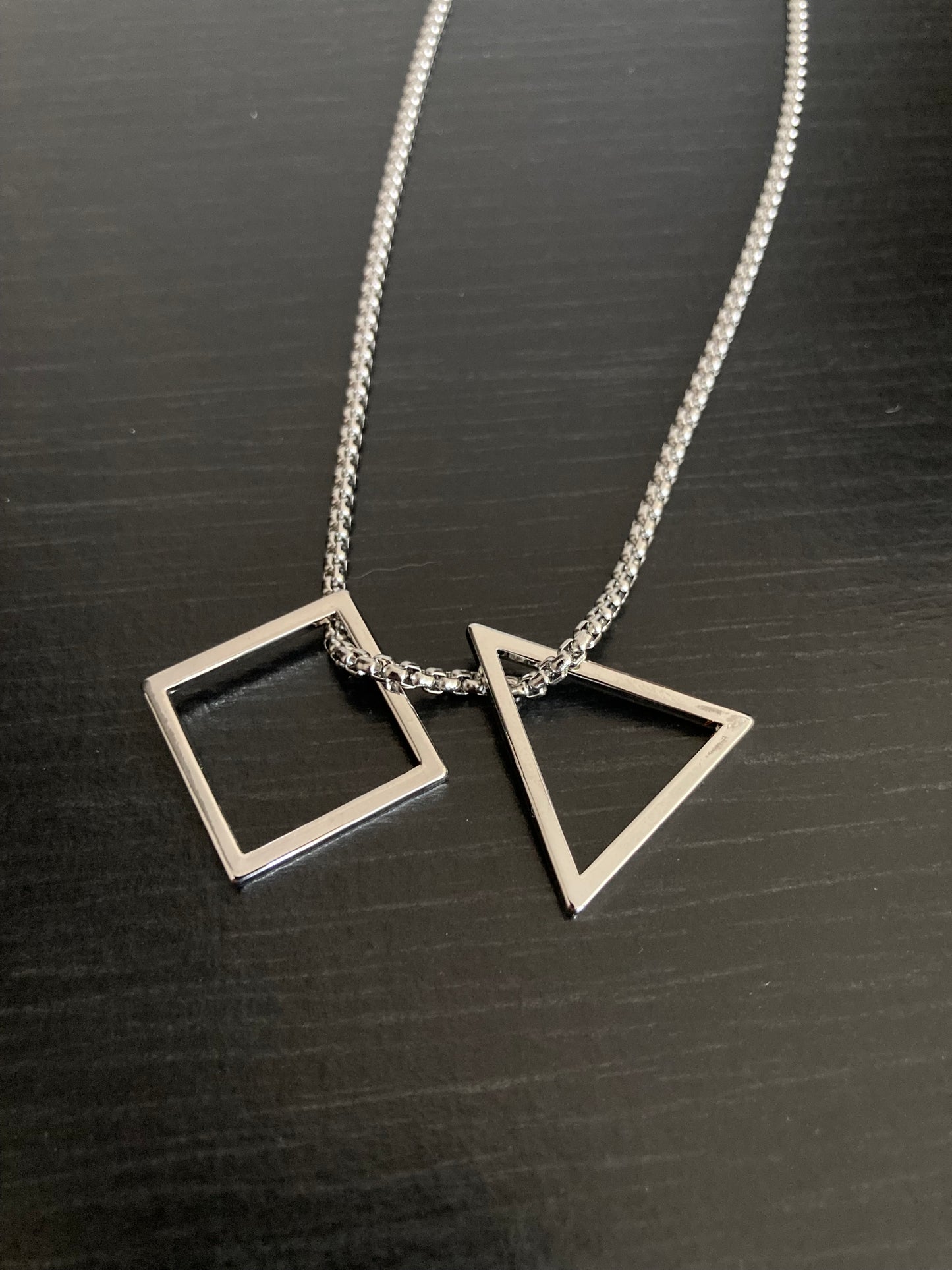 Lying on a black surfacec are a triangle and square pendant. These are seperate items on a chain and both are hollow. Silver in colour and made of stainless steel these are both shiny and high quality.