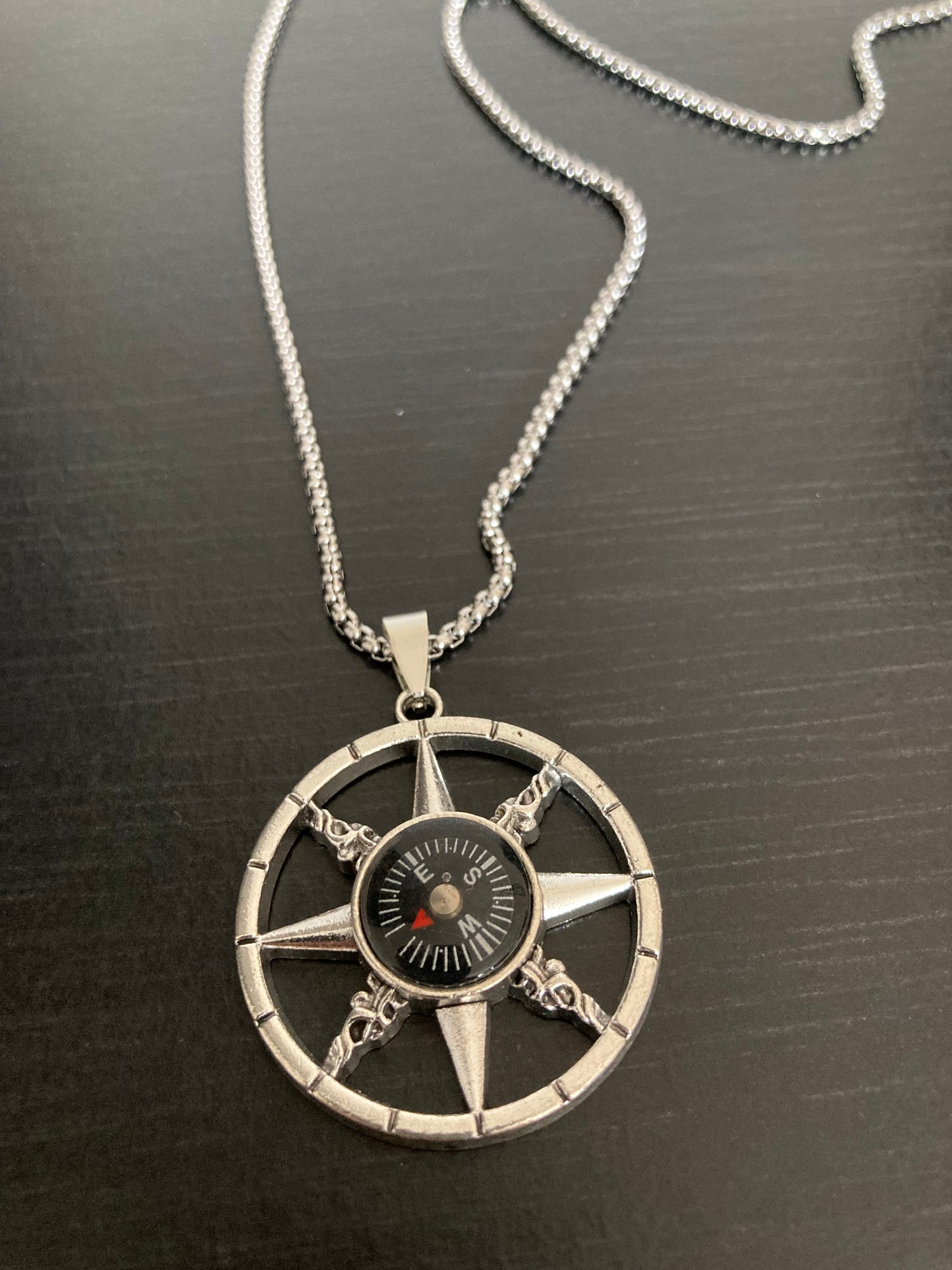 A view from above of a fully working compass necklace. Attached to a stainless steel chain the pendant is a circle shape with an outer ring which has eight cut out points leading from there to the centre where the compass sits.