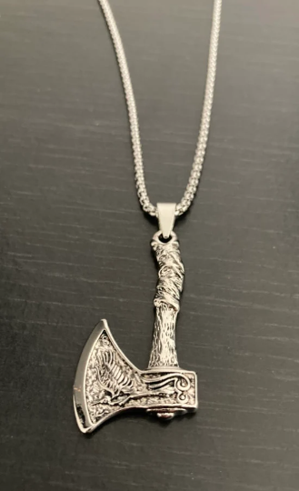 On a black surface is a stainless steel silver colured necklace. The pendant is in the shape of an axe with a design of a raven on the heaqd of it. You can see the chain which is the same material and colour and see how shiny this whole item is.