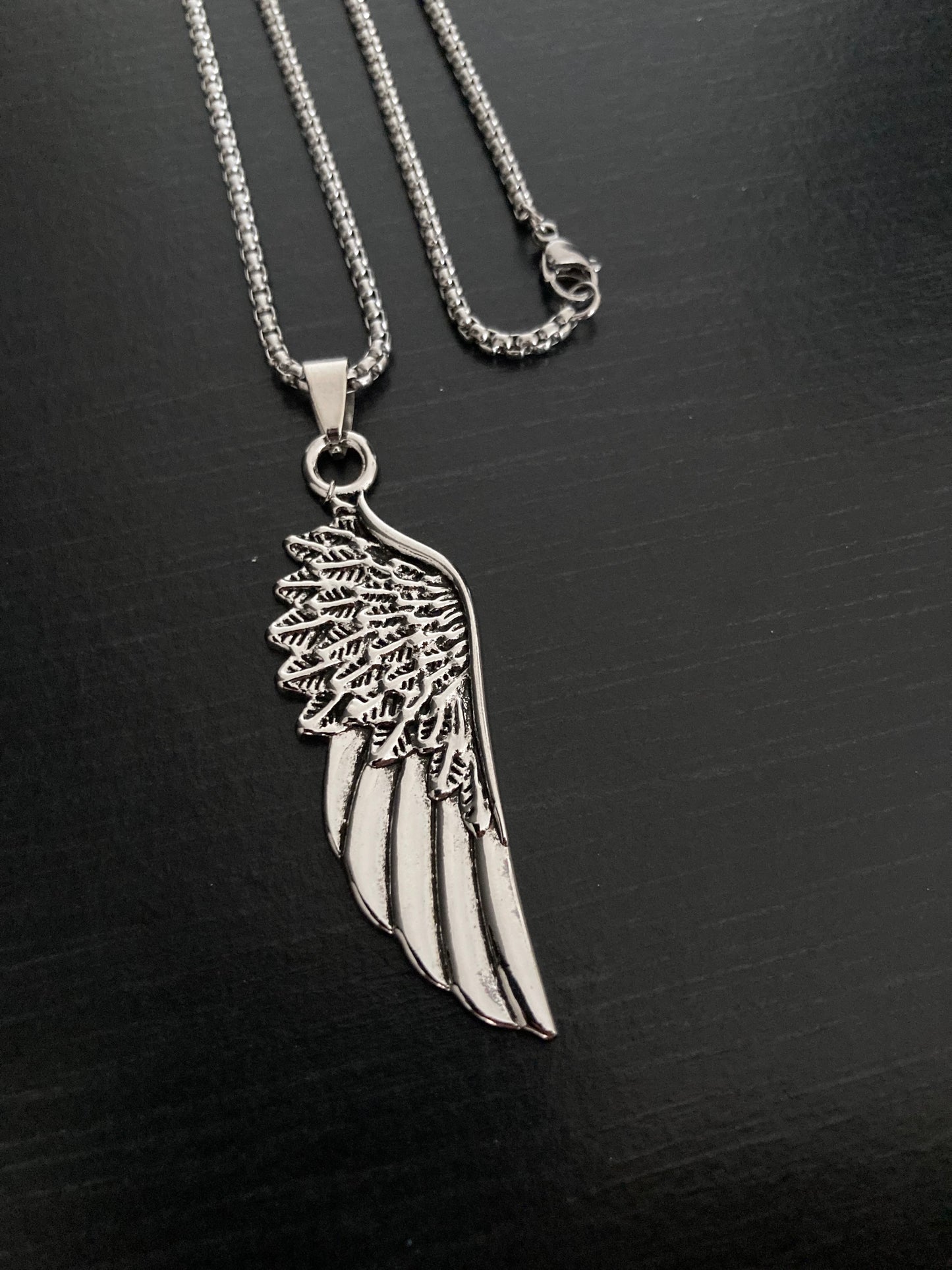 A solid looking chain with a clasp for fasterning are sitting on a black backgrlound. Attached to this is a pendant in the shape of a wing which is silver in colour. The engravings on this are made to look like feathers and ar very detailed.