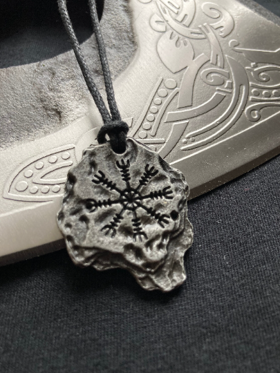 Leaning against a silver axe blade is a black corded necklace featuring a pendant in the shape of a jagged rock with a helm of awe inscription on it. The item is shiny but also has the appearance of a rock so no defined shape to it and is solid.