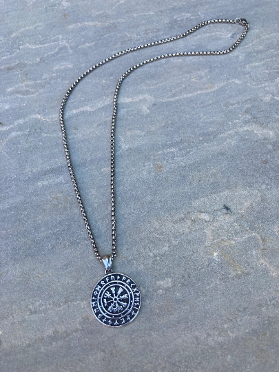 The vegvisir necklace is lying on a grey textured stone background with the full length of it on display. The chain looks and feels weighty and is silver in colour. The main feature of the item is black with silver carvings of runes and a compass.