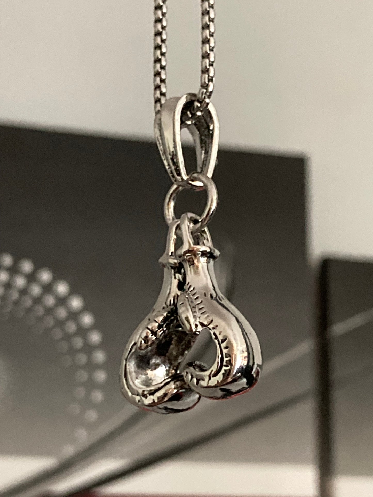 A silver coloured chain is dangling with a pendant attached to it which is of a pair of boxing gloves. These are facing each other so the tips at the bottom touch. With detailed engraved markings they look and feel like a quality item. 